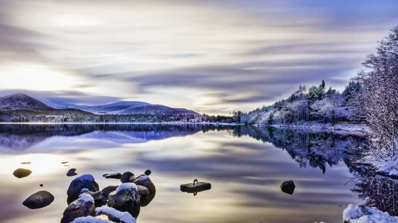 Beautiful winters day with soft clouds, snow on trees and rocks, reflections on calm water in Scotland in January.