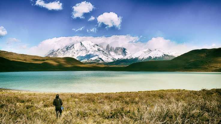 A trekker stands on a lakeshore and looks at view of snow-covered mountains